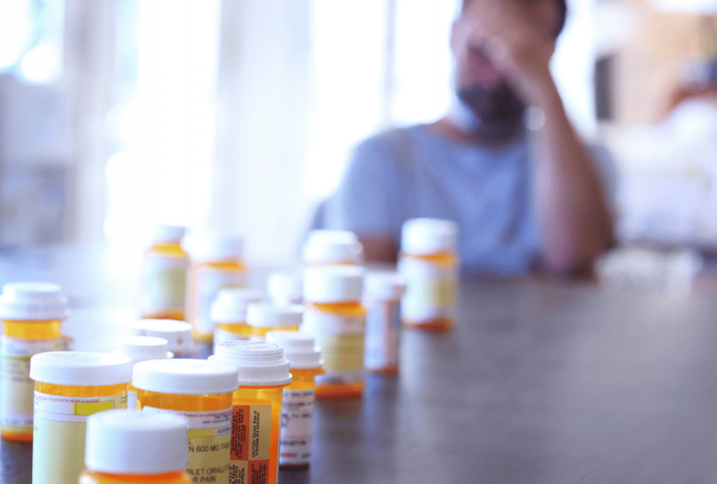 man sitting at a table with his head in his hand behind a row of prescription bottles